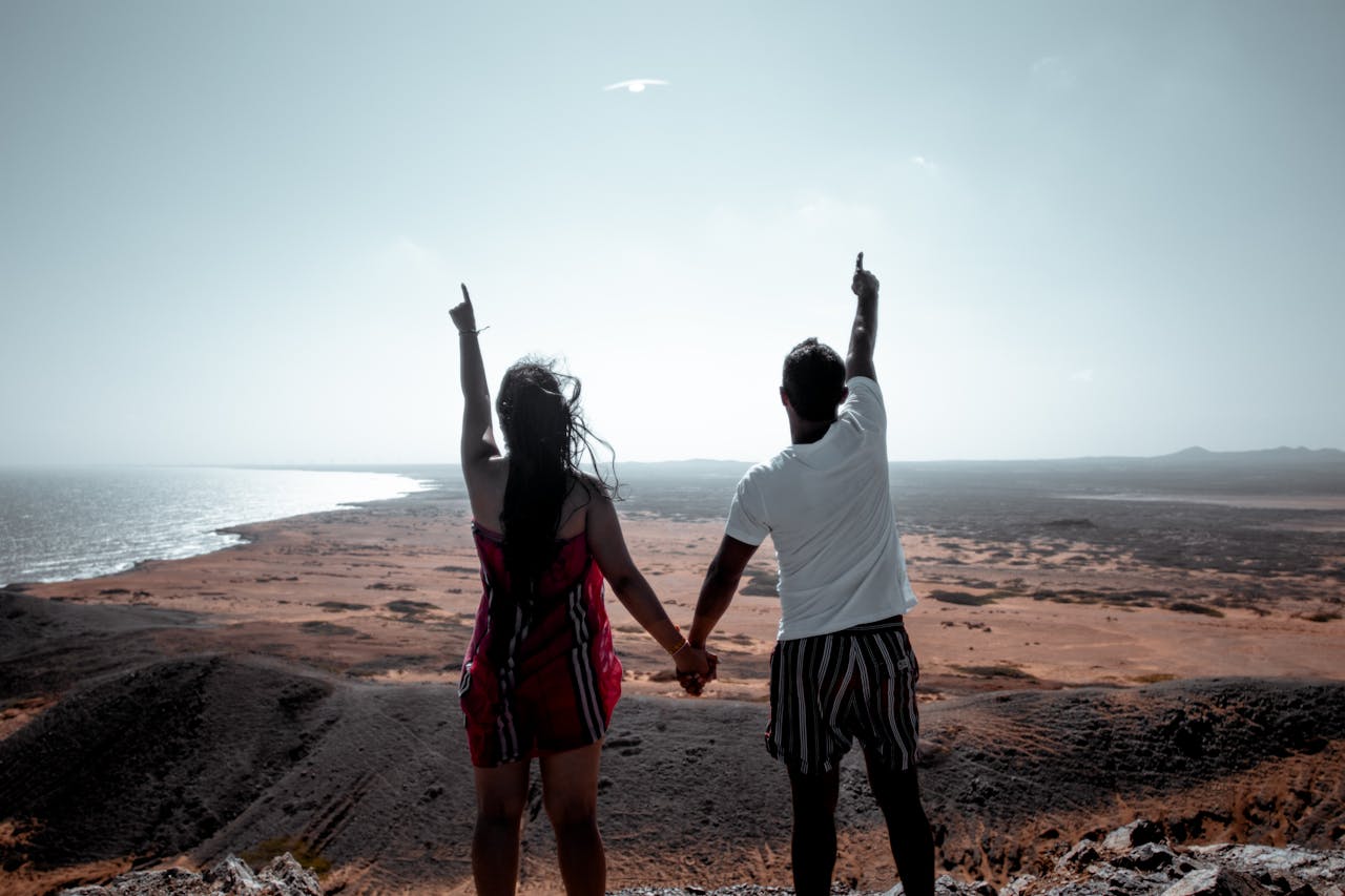 A couple holding hands and pointing at the horizon, enjoying a scenic sunset view by the beach.